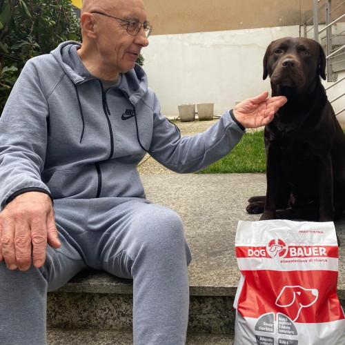 Uomo accovacciato con due cani Boxer e sullo sfondo sacco di crocchette Dogbauer